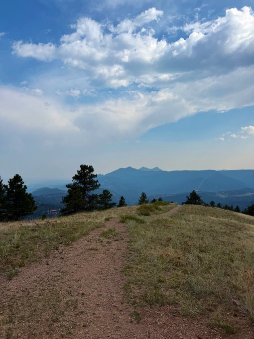 A scenic mountain landscape with a dirt trail winding through grassy terrain toward distant blue mountain ranges under a partly cloudy sky.