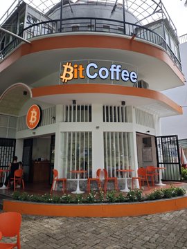 The image shows the storefront of "₿itCoffee," a café featuring its Bitcoin-inspired logo prominently displayed above the entrance, with outdoor orange seating and potted flowers. The post appears to thank a visitor named @timbo_xyz for stopping by ₿itCoffee and references the Coldcard hardware wallet brand, with the Bitcoin symbol (₿) in the name tying into the cryptocurrency theme.