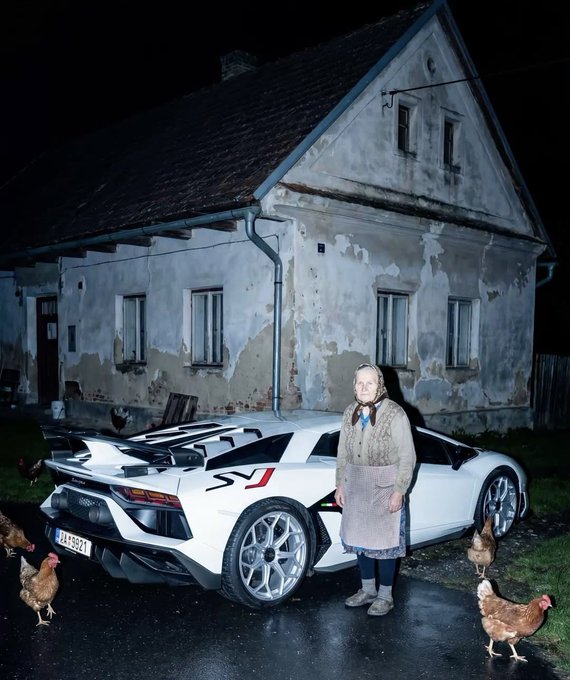 An elderly woman in traditional attire stands in front of a white Lamborghini with "SV" branding, parked outside an old weathered house with chickens roaming around.