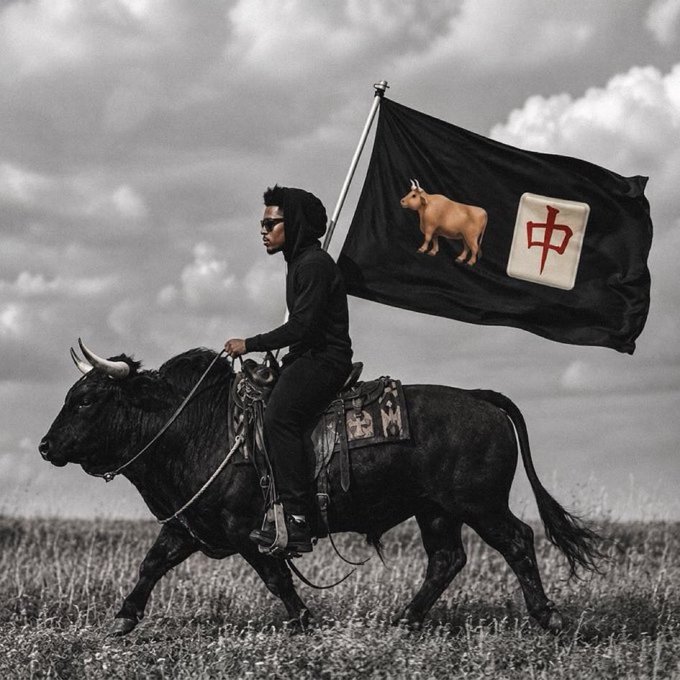 A black and white edited photo of a man riding a black bull while holding a flag featuring a bull and a mahjong tile with the Chinese character 中 (zhong).