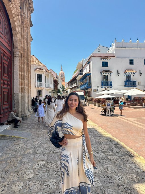 A woman smiling in a white and blue patterned outfit standing on a cobblestone street in what appears to be a historic town with white colonial-style buildings and a church in the background.