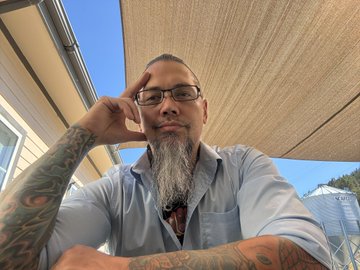 A man with a gray beard, glasses, and heavily tattooed arms rests his head on his hand under a tan awning, posing contemplatively for the selfie. The image humorously pairs with the caption "this is me / this is my ape," as his bearded, thoughtful expression makes the playful comparison fitting.