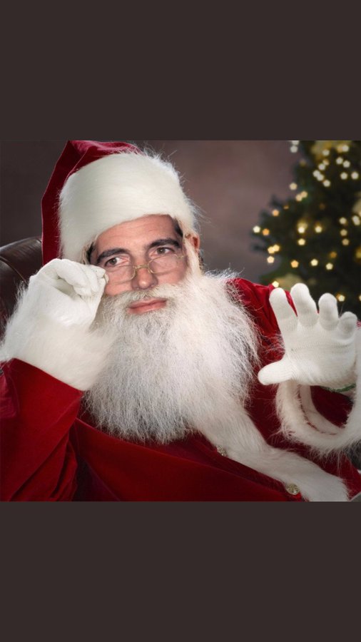 A man dressed as Santa Claus with a white beard and glasses adjusts his spectacles with a gloved hand, posed next to a lit Christmas tree.
