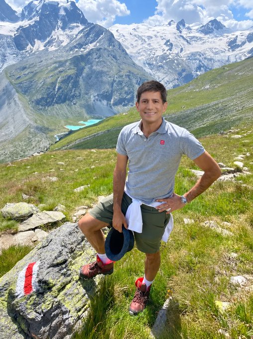 A man wearing a grey polo shirt and olive green shorts stands smiling in a mountainous alpine landscape, holding a cap with a white sweatshirt tied around his waist, with snow-capped peaks and a turquoise lake in the background.