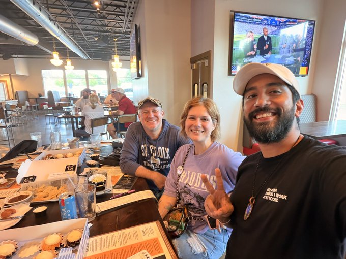 Three people smile for a selfie at a restaurant table with food and drinks, one wearing a 'St. Croix' shirt, one a 'Mudd' shirt, and one a black 'Intuit Work Hard Play Hard' shirt.
