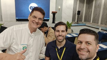 A selfie photo of three men, one older man in a white shirt smiling broadly on the left, and two younger men, taken indoors at what appears to be a conference or event.