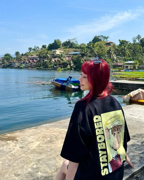 A woman with red hair wearing a black "STAYBORED" t-shirt featuring the Bored Ape Yacht Club logo stands on a dock by the water, promoting the brand's free merchandise giveaway alongside traditional boats in a tropical harbor setting.
