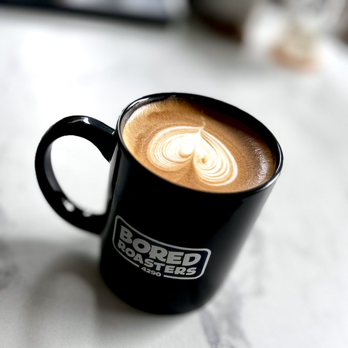 A black ceramic mug with the text 'BORED ROASTERS 4290' sits on a marble countertop, filled with a coffee drink featuring heart-shaped latte art.