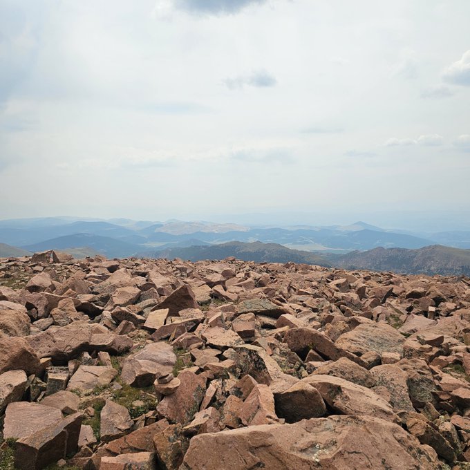 Mountain summit landscape photograph showing a foreground of reddish-brown boulders with rolling mountain ranges and a cloudy sky in the distance.