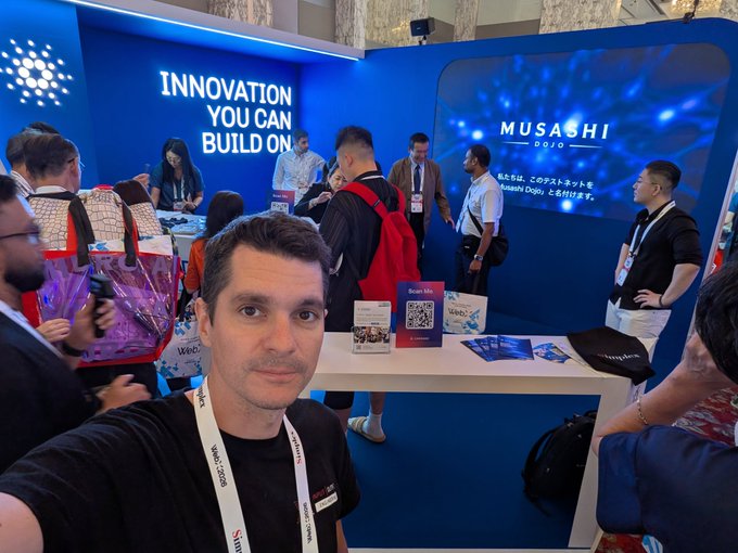 A man takes a selfie at a tech conference booth displaying 'INNOVATION YOU CAN BUILD ON' and 'MUSASHI DOJO' with Japanese text, surrounded by attendees and promotional materials.