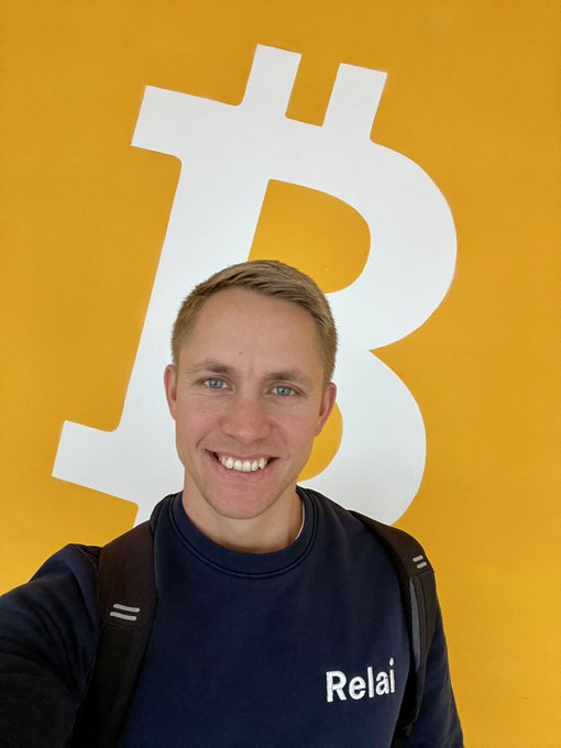 A selfie of a smiling man wearing a dark blue sweatshirt with 'Relai' printed on it, standing in front of a yellow wall with a large white Bitcoin logo.