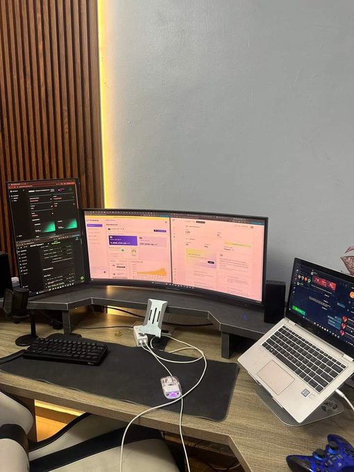 A desk setup featuring a vertical monitor, two horizontal monitors, and a laptop displaying trading charts, alongside a phone mount on a wooden slat wall.
