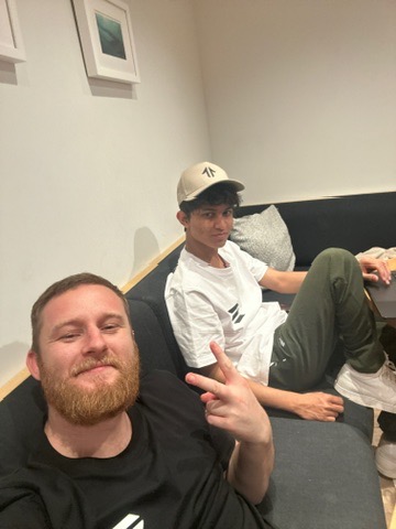 Two guys taking a selfie—one in a black shirt and another in a white t-shirt and NY Yankees cap—sitting on a couch together. The image captures the early grind mentioned in the caption, showing the humble beginnings and camaraderie between the two co-founders before their project's success.