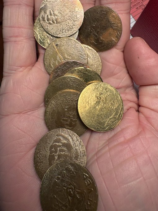 A hand holds several worn, gold-colored coins, likely depicting Buddhist temples or shrines, that appear to be Japanese temple omamori or amulets.