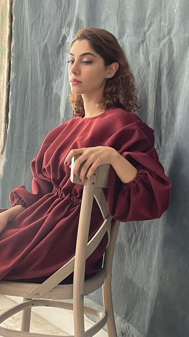 The image features a woman sitting on a wooden chair, wearing a burgundy dress with a high neckline. She has her hands resting on her lap while looking off into the distance. The background of the photo appears to be a gray curtain or wall, which contrasts with the vibrant red color of the woman's dress and adds depth to the image. The post seems to be promoting an event or gathering related to tezostuesdays, as indicated by the caption "Happy Tezostewsday !!!".