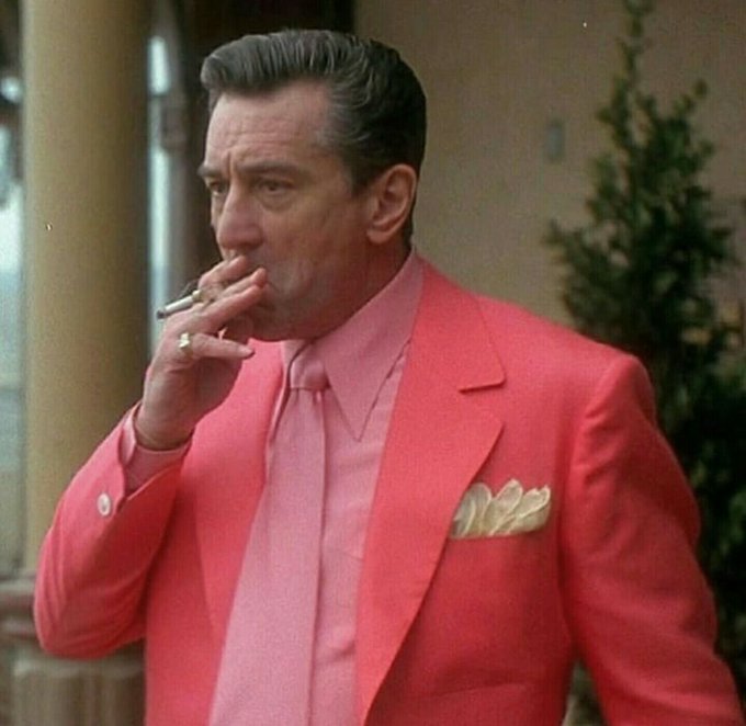 A man in a bright pink suit and matching shirt and tie, smoking a cigarette outdoors.
