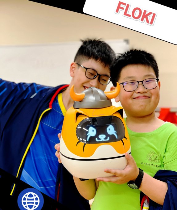 Two smiling boys pose together while holding a white and orange robot with a horned helmet, with a 'FLOKI' label visible in the top right corner.