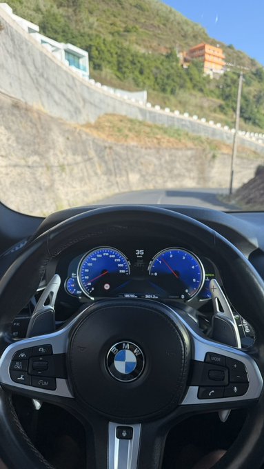 A driver's POV shot from behind the wheel of a BMW, showing the digital dashboard with a speedometer reading 35 mph and the iconic BMW logo on the steering wheel, as the car travels along a scenic mountain road. The post jokingly ties in the "GM" (good morning) greeting with a request for crypto tips on the Solana blockchain, using the BMW's "BMW" branding as a playful nod to the community vibe.