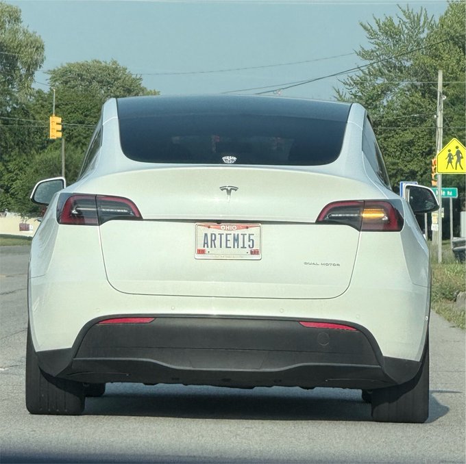 The rear view of a white Tesla Model Y driving on a road, displaying an Ohio license plate that reads 'ARTEMI5' with a small decal of cherries above it.