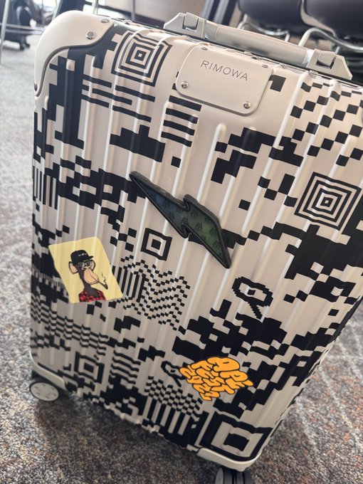 A Rimowa suitcase covered in a black and white pixelated digital glitch pattern, featuring several stickers including a cartoon monkey and an orange bee logo, with an airplane-shaped sticker visible—tying into RTFKT's cryptobro aesthetic of treating travel as another on-chain flex.