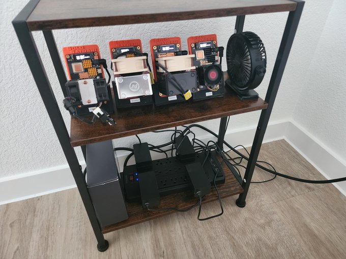 Several Bitmain mining units sit on a shelving rack alongside cooling fans and a power distribution system, with tangled cables below. The setup reflects the post’s urgency to get the $BTC miners running in the new house.