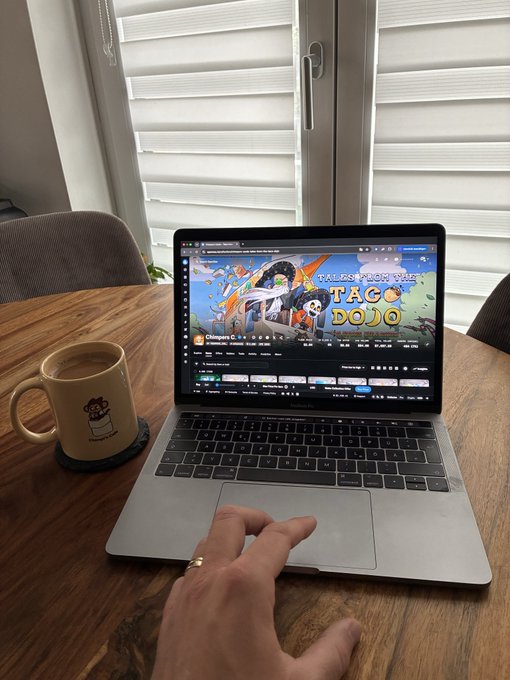 A person's hand using the trackpad of a MacBook on a wooden table next to a mug, with a YouTube video titled 'TALES FROM THE TACO DOJO' playing on the screen.
