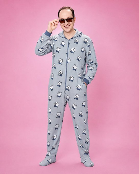 A man wearing sunglasses and a gray onesie pajama covered in a repeating cartoon sheep pattern, posing against a pink background.