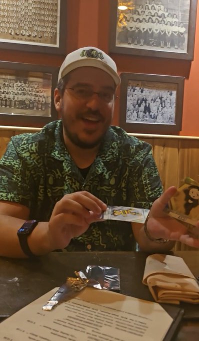 A man wearing a green patterned shirt and a white cap with an emblem, sitting at a table and holding what appears to be a Pokémon card, with a menu and other items on the table in front of him.
