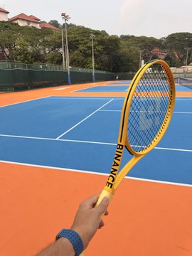 A first-person view of a hand holding a yellow tennis racket over a blue and orange tennis court, with houses and trees visible in the background.