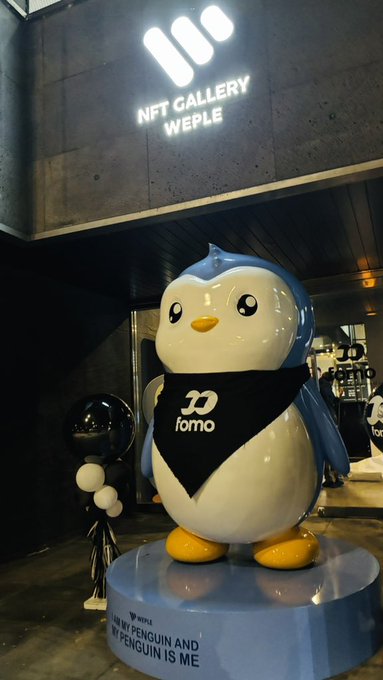 A large penguin mascot statue wearing a black bandana with the 'forno' logo stands on a circular blue platform in front of an 'NFT GALLERY WEPLE' storefront with an illuminated logo above.
