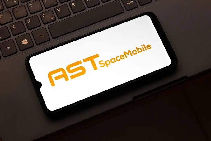 A smartphone resting on a black laptop keyboard displays the orange AST SpaceMobile logo on a white background.