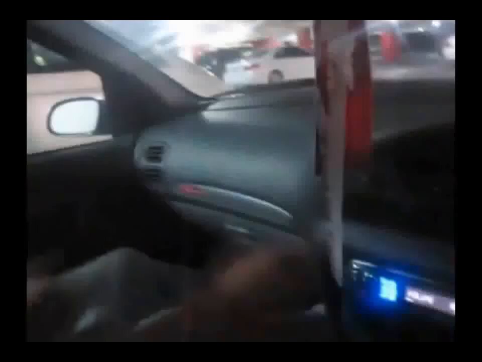 The image shows a car interior with a person's hand visible inside the vehicle. The background of the image features a parking lot filled with cars. There are multiple cars parked at various positions, including one that appears to be a red car. A sign in the parking lot reads "Micky: $TIBBIR holders".