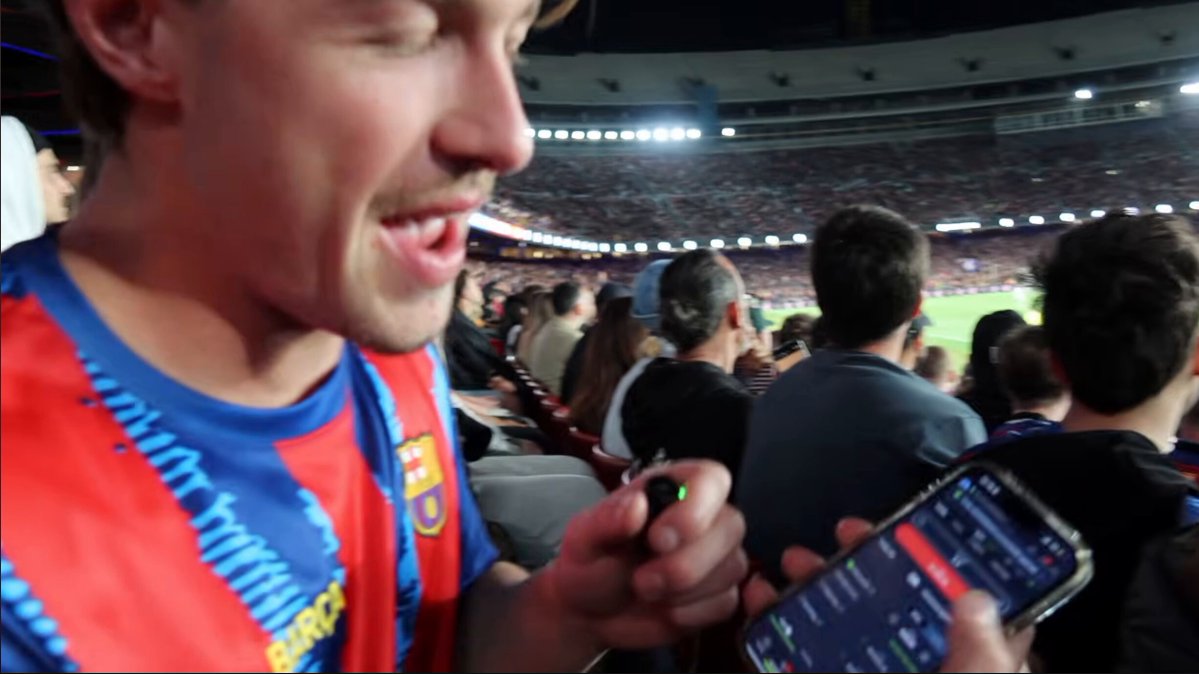 ?"What's happening here? A man holding a smartphone showing its screen which displays a cryptocurrency page. The context behind this scene involves people attending a large stadium event, possibly watching a sports game or an important live event where they might discuss Bitcoin or similar topics. In addition, there are several other individuals present in the image who could be engaged in conversations about Bitcoin and related subjects."