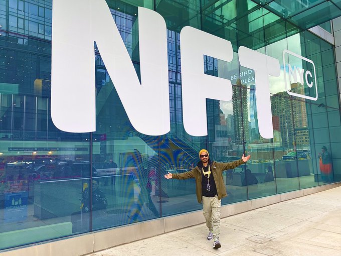 A person wearing a yellow beanie and brown jacket poses with arms outstretched in front of a large glass building displaying 'NFT' and 'BEYOND PLE... NYC' signage.