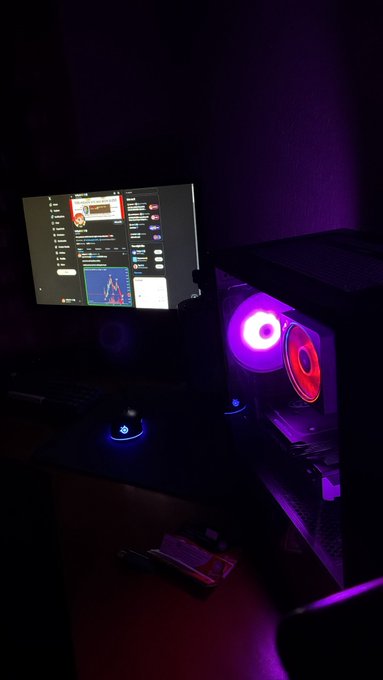 A dimly lit gaming setup featuring a monitor displaying a social media feed, a PC case with glowing purple and red RGB fans, and a mouse on an illuminated mousepad.