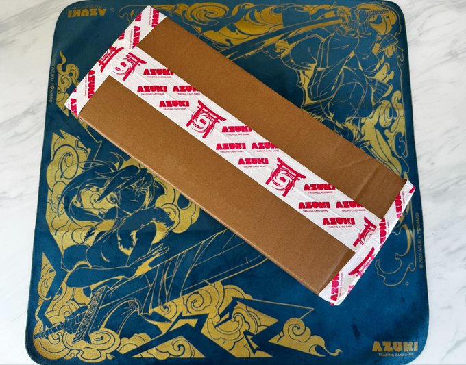 A long cardboard shipping box sealed with red and white 'AZUKI TRADING CARD GAME' branded tape rests on top of a dark blue and gold playmat featuring anime-style character artwork.
