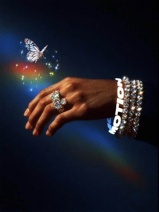 A stylized artistic image showing an outstretched hand adorned with rings and a rhinestone bracelet, with a glowing rainbow trail of sparkles and a monarch butterfly, featuring the text 'IN MOTION'.
