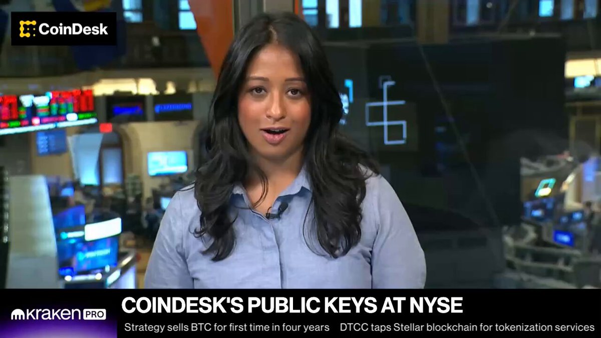 In the image, a woman wearing a blue shirt is standing in front of a computer screen displaying an article about CoinDesk's public keys at the NYSE. The article talks about the importance of these keys for cryptocurrency exchanges like theirs. The woman appears to be engaged with the content and possibly discussing it with someone else or taking notes on her phone.