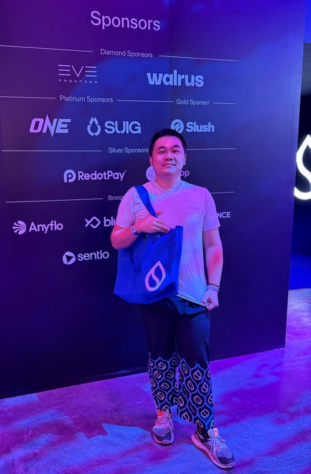 A man posing in front of a Sponsors wall at an event, holding a blue Sui-branded bag, with logos including Walrus, ONE, Slush, RedotPay, Anyflo, and Sentio visible.