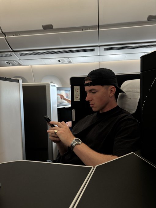 A man in a black cap and t-shirt sits in an airplane seat, looking at his smartphone.