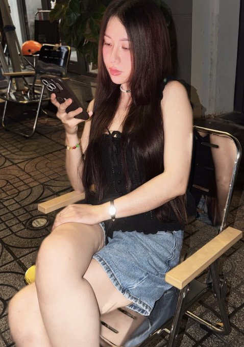 A young woman with long dark hair wearing a black corset top and denim skirt sits on a folding chair while looking at her smartphone.