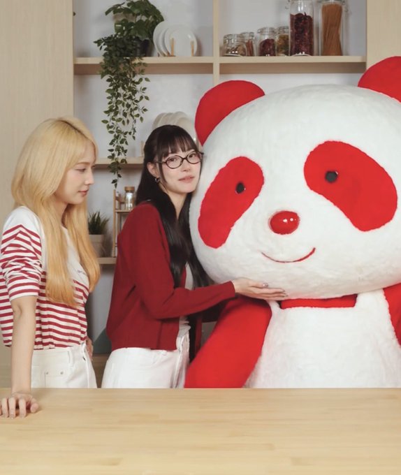 Two young women pose next to a giant plush red and white panda mascot in a room with shelves of plants and glass jars in the background.