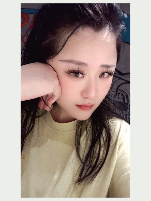 A close-up selfie of a young woman with long dark hair, wearing a pale yellow shirt and posing with her hand near her face.