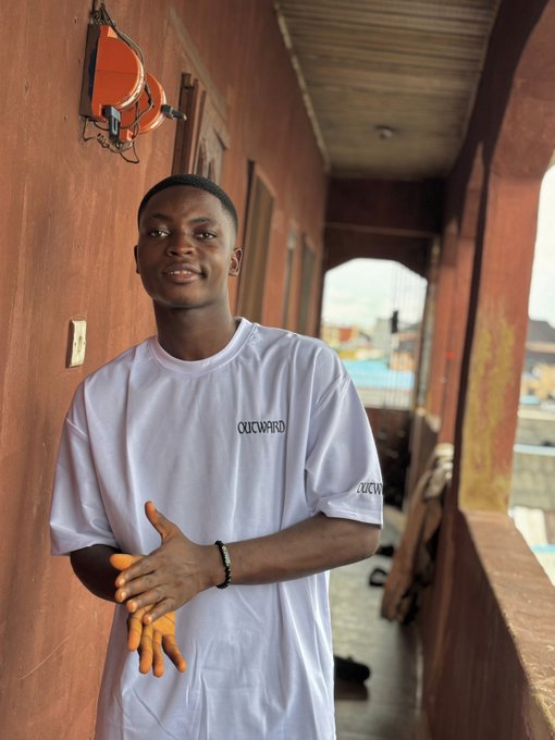 A young man smiling at the camera, wearing a white t-shirt with "OUCHWARD" printed on the chest, standing on a balcony with an orange-brown wall behind him.