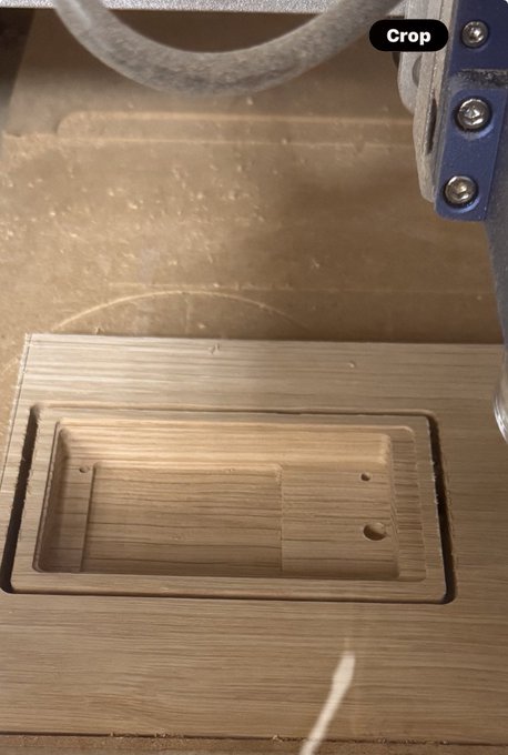 Close-up of a CNC milling machine carving a rectangular cavity with a small circular hole into a piece of wood, with a 'Crop' watermark visible at the top.
