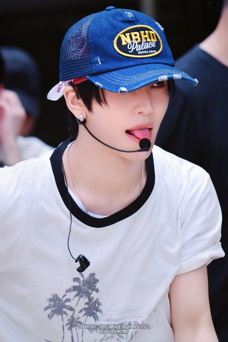 A young performer with dark hair wears a distressed blue NBHD Palace cap, white t-shirt with palm tree print, and headset microphone while singing during a stage performance. The watermark credits "NEUZMADE RIGHTS RESERVED" and "LENORMKIN ONG," with the post identifying the performer as Xin Long (씬롱/鑫隆/シンロン), a member of AlphaDriveOne (ALD1).