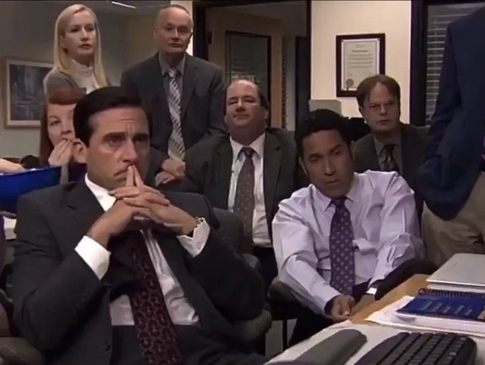 A scene from the TV show The Office showing Michael Scott and several coworkers sitting in an office setting watching a presentation attentively.