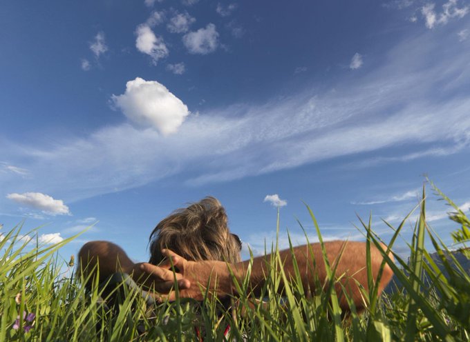 A low-angle shot of a person lying in tall green grass, looking up at a bright blue sky dotted with fluffy white clouds.