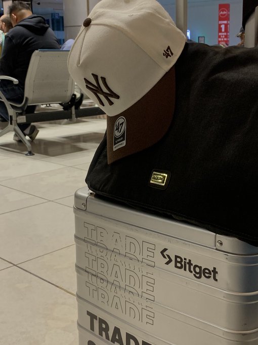 A cream and brown "47" New York Yankees cap rests atop a black bag and a silver suitcase branded with "TRADE" and the "Bitget" logo, signaling a return from travel to engage with the Bitget community. The image visually ties to the caption by marking the transition from a weekend getaway back to the travel-ready "natural habitat" of trading and building with @BitgetFans_.