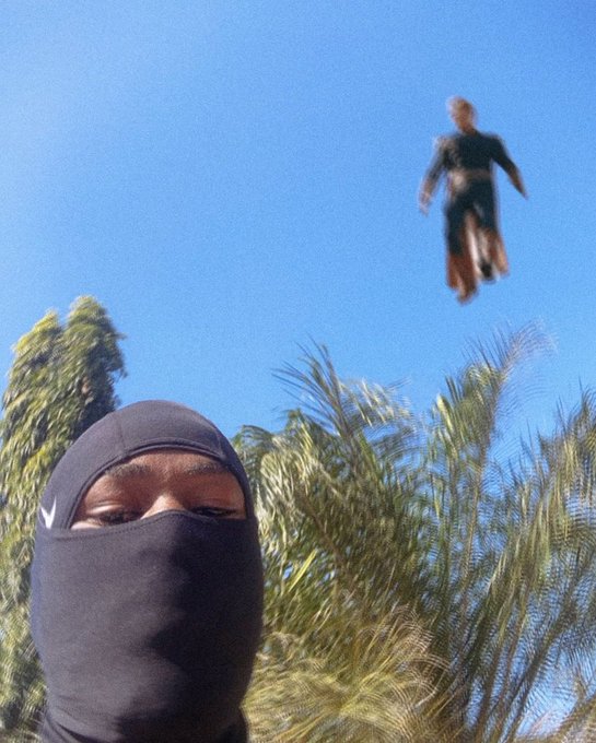 In the foreground, a person wears a black balaclava, set against a vibrant blue sky with foliage below. Above, a figure in a black suit is seemingly levitating, echoing the adventurous spirit conveyed by the social media post, "gm, it's midweek, never run out of motion," suggesting a metaphorical or literal ascent at the week's midpoint.